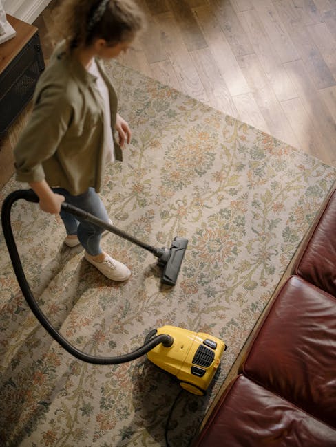 A professional cleaner wearing a black glove is using a spray wand connected to a cleaning machine to deep clean and sanitise a textured, dark grey carpeted floor in a commercial or residential settin